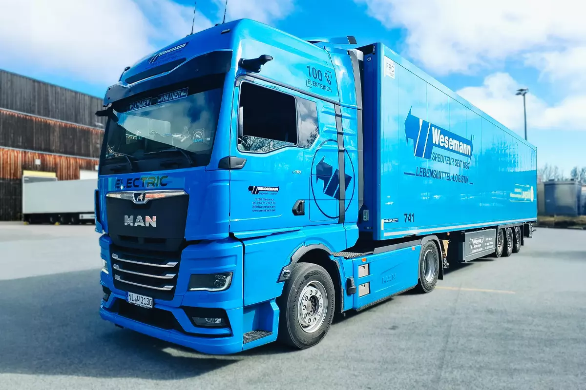 Blauer elektrischer LKW der Spedition Wesemann mit Kühlaufbau steht auf einem Betriebshof unter blauem Himmel. Auf dem Fahrerhaus ist der Schriftzug „100 % elektrisch“ zu sehen, das Fahrzeug trägt das Wesemann Logo und steht für nachhaltige Lebensmittellogistik.